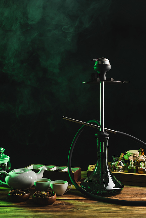 Hookah near cups and sty tea on wooden surface on black background with smoke