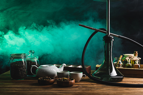 Hookah, dry tea and teapot on wooden surface on black background with smoke