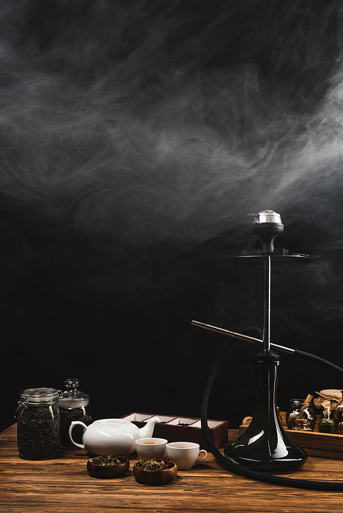 Cups of tea, hookah on wooden surface on black background with smoke