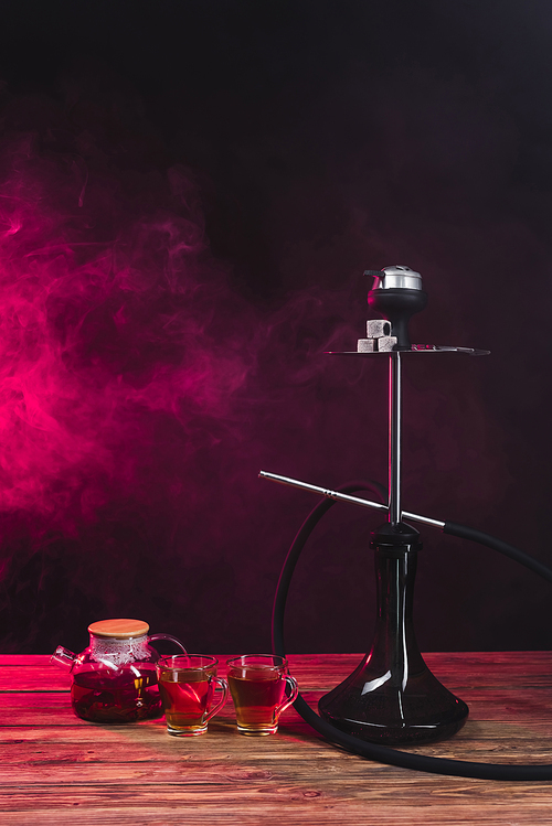 Hookah near cups of tea on wooden surface on black background with pink smoke