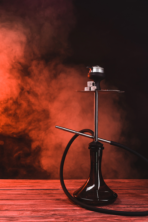 Hookah with coals on wooden surface on black background with red smoke