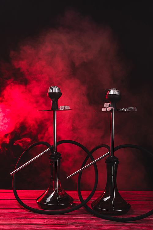 Hookahs with coals on wooden surface on black background with red smoke