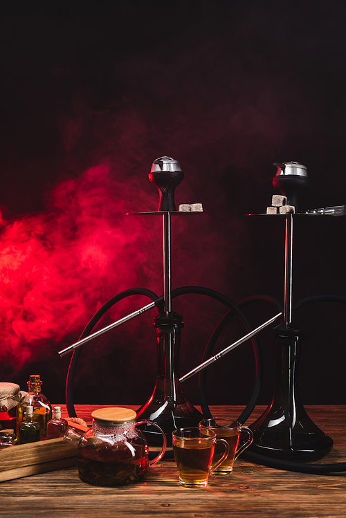 Cups of tea near teapot and hookahs on wooden surface on black background with red smoke