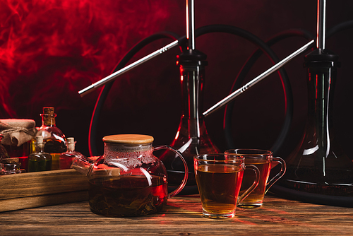 Tea near hookahs on wooden surface on black background with smoke