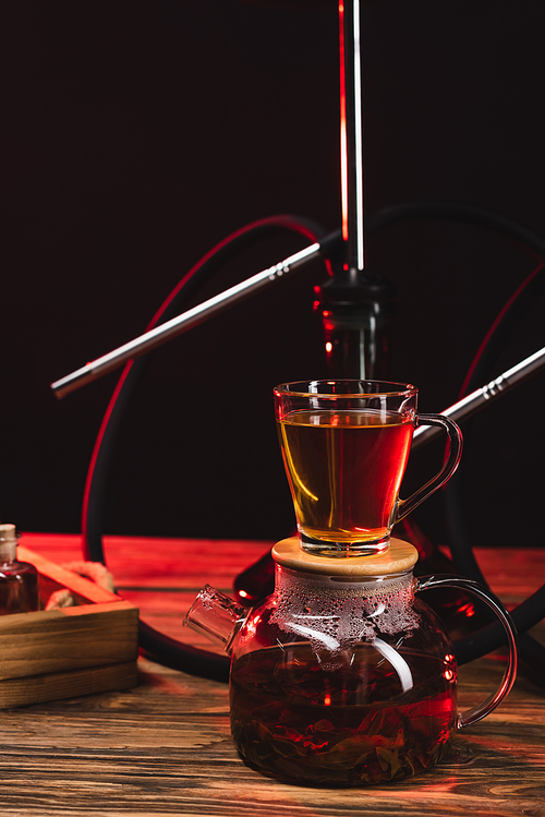 Cup of tea on teapot near hookah on wooden surface isolated on black