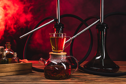 Cup of tea on pot near hookahs on wooden surface on black background with red smoke