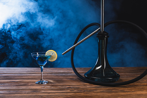 Glass with cocktail near hookah on wooden surface on black background with smoke