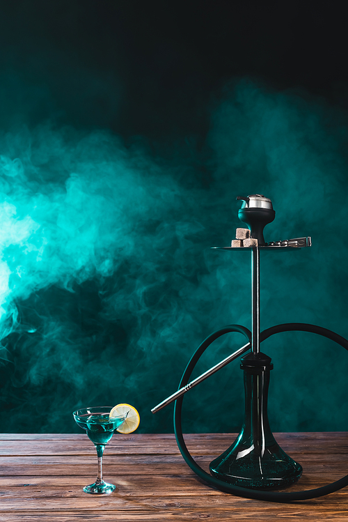 Hookah near glass of cocktail on wooden surface on black background with green smoke