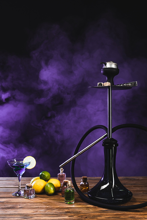 Hookah near cocktail on wooden surface on black background with purple smoke