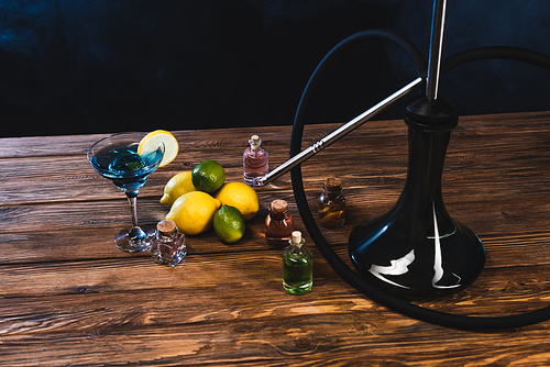 Glass of cocktail near lemons and hookah on wooden surface on black background with smoke