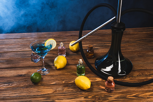 Hookah near lemons and glass of cocktail on wooden surface on black background with blue smoke