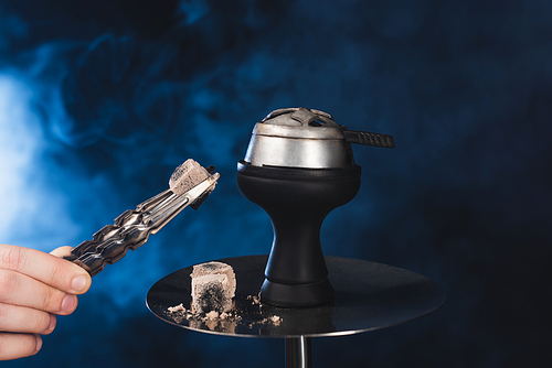 Cropped view of man holding tweezers with coal near hookah on black background with smoke