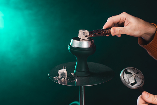 Cropped view of man holding coal in tweezers near hookah on green background with smoke