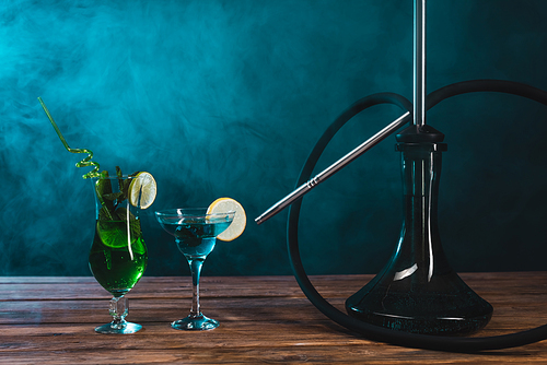 Glass of cocktails near hookah on wooden surface on black background with green smoke