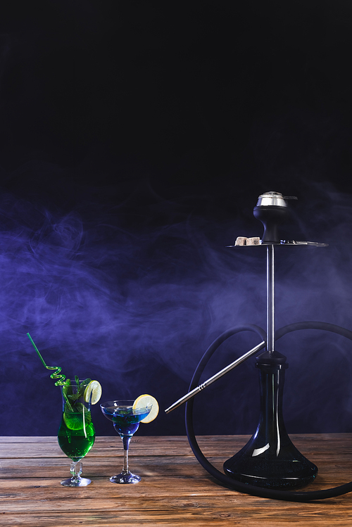 Hookah near glasses of cocktails on wooden surface on black background with smoke