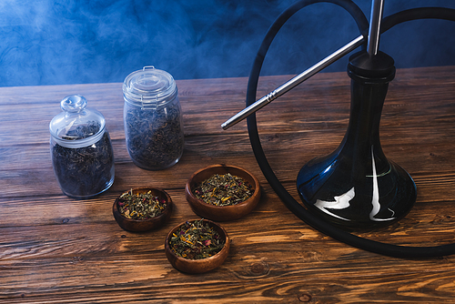 Dry tea in jars and bowls near hookah on wooden surface on black background with smoke