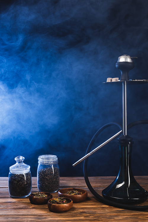 Hookah near dry tea in bowls on wooden surface on black background with blue smoke