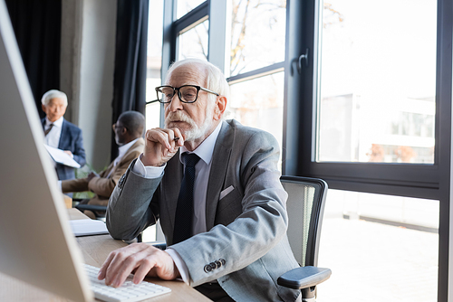 senior businessman in eyeglasses working on computer near blurred multicultural colleagues