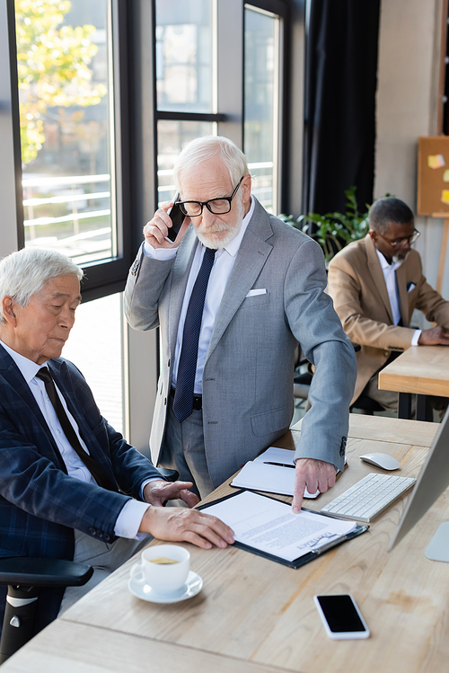 senior businessman pointing at contract near asian colleague while talking on mobile phone