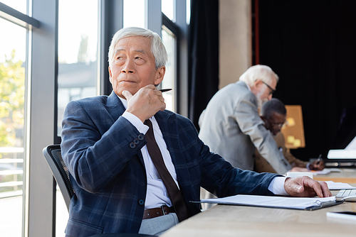 senior asian businessman looking away while working near blurred multiethnic colleagues