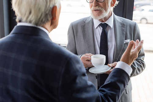 cropped view of senior businessman with coffee cup near blurred colleague pointing with hand