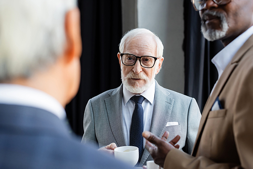 elderly businessman in eyeglasses holding coffee cup near blurred interracial colleagues