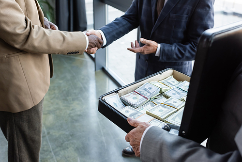 partial view of interracial business colleagues shaking hands near man holding briefcase with money
