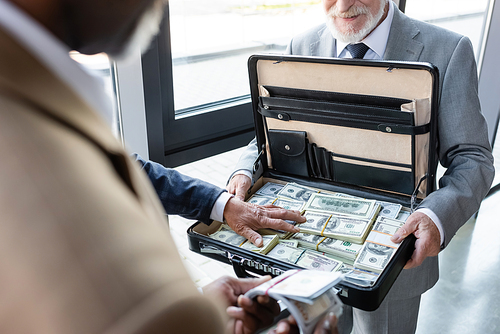 partial view of senior businessman holding briefcase with money near blurred interracial business partners