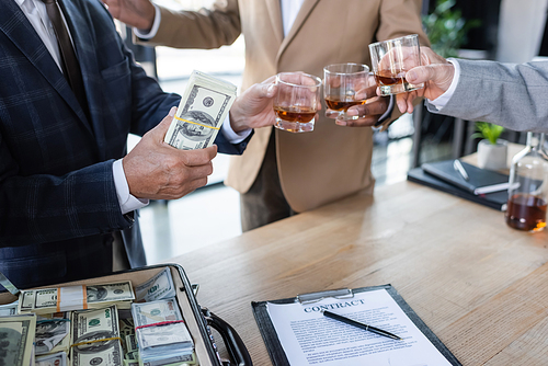 cropped view of businessman holding dollars while clinking glasses near contract and briefcase