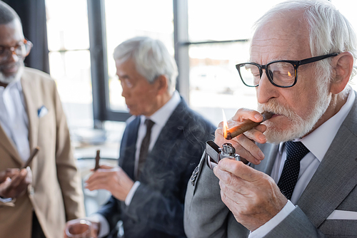 senior successful businessman lighting cigar near blurred multiethnic business partners talking in office