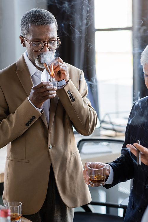 african american businessman in eyeglasses lighting cigar near colleague with glass of whiskey