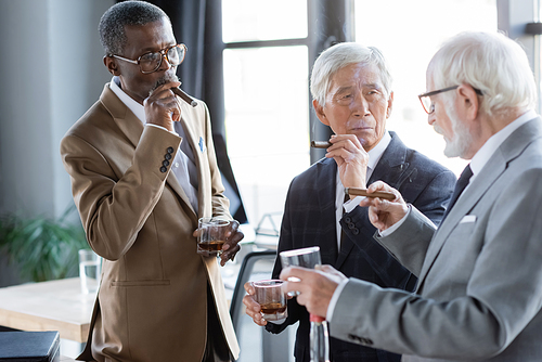 elderly multiethnic business people with cigars and glasses of whiskey talking in office