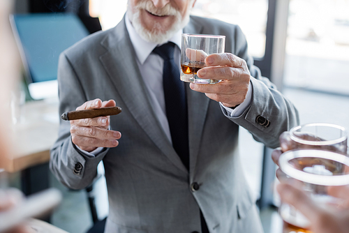 cropped view of elderly businessman holding cigar and glass of whiskey near blurred colleagues