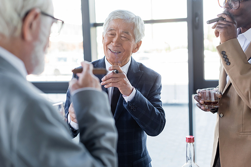 elderly asian businessman smiling near interracial colleagues smoking cigars and drinking whiskey in office