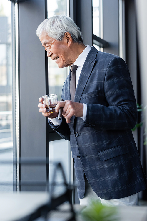 senior asian businessman with whiskey and cigar smiling while looking down through office window