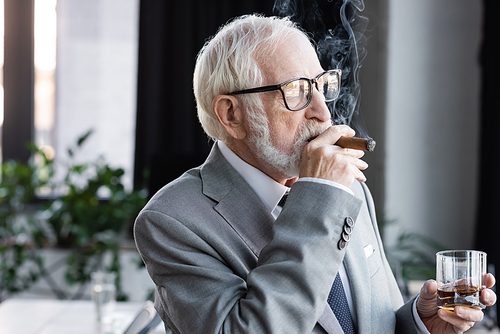 senior businessman with glass of whiskey looking away while smoking cigar in office