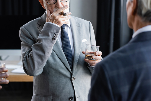 partial view of senior businessman with glass of whiskey smoking cigar near blurred interracial business partners