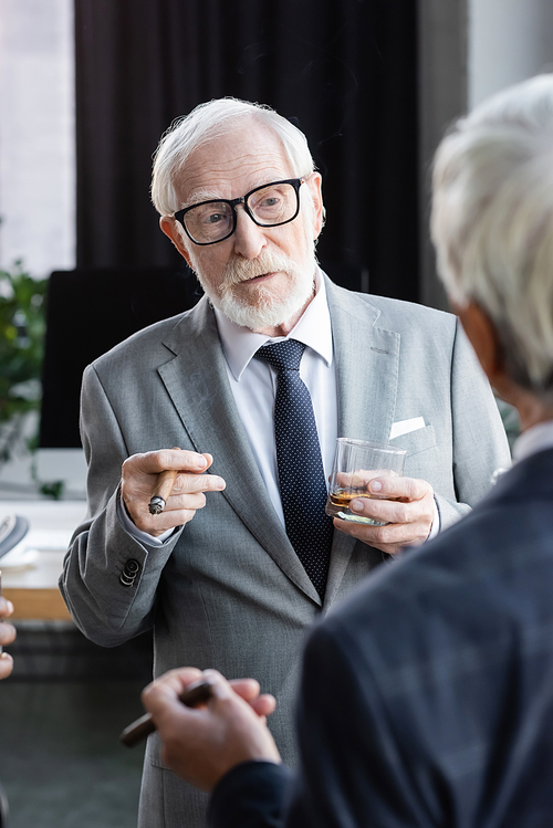 elderly businessman in eyeglasses holding cigar and glass of whiskey near colleague on blurred foreground