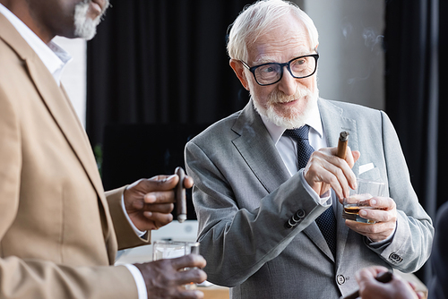 senior businessman with cigar and whiskey looking away during discussion with blurred african american colleague