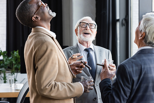 senior multiethnic businessmen laughing while drinking whiskey and smoking cigars in office