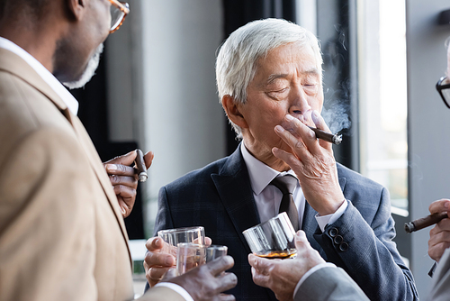 senior asian businessman smoking cigar while clinking glasses of whiskey with blurred interracial colleagues