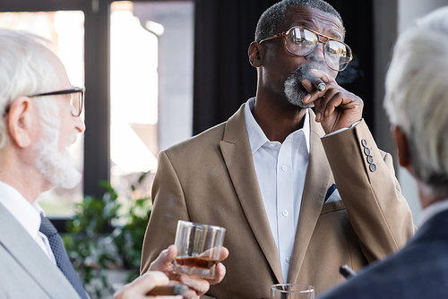 senior african american businessman in eyeglasses smoking cigar near blurred colleagues  with glasses of whiskey
