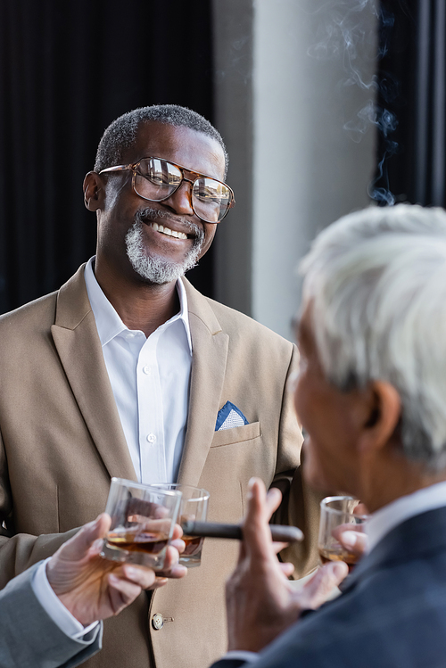 cheerful african american businessman in eyeglasses standing with cigar and glass of whiskey near blurred senior colleagues