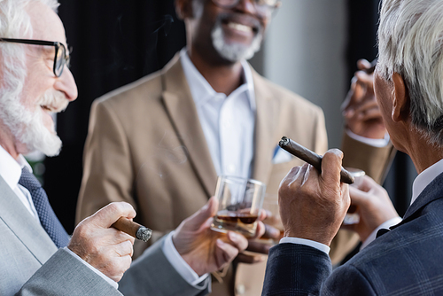 senior businessman holding glass of whiskey and cigar near smiling multiethnic colleagues