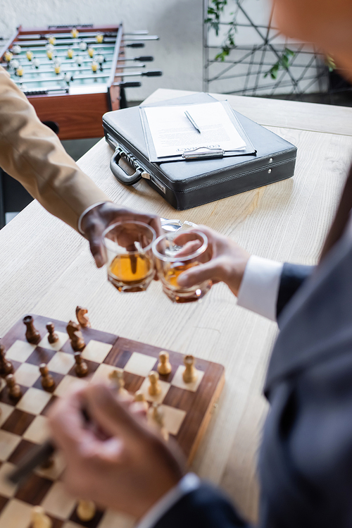 cropped view of blurred business partners clinking glasses near chessboard and contract on briefcase