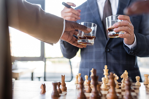 cropped view of successful interracial business partners holding glasses of whiskey above blurred chessboard