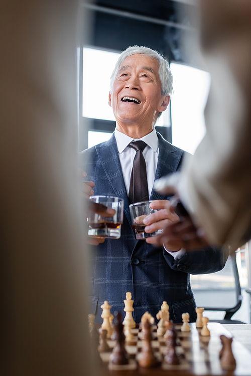 senior asian businessman smiling near chessboard and blurred colleagues with glasses of whiskey