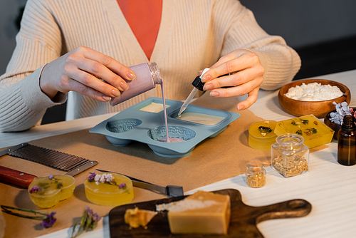 Cropped view of craftswoman pouring soap in mold near flowers, cutter and flakes on table