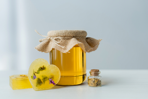 Jar with honey near handmade soap bars and herbs on table on grey background