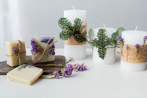 Craft soap bars with flowers on cutting board near candles on table on grey background